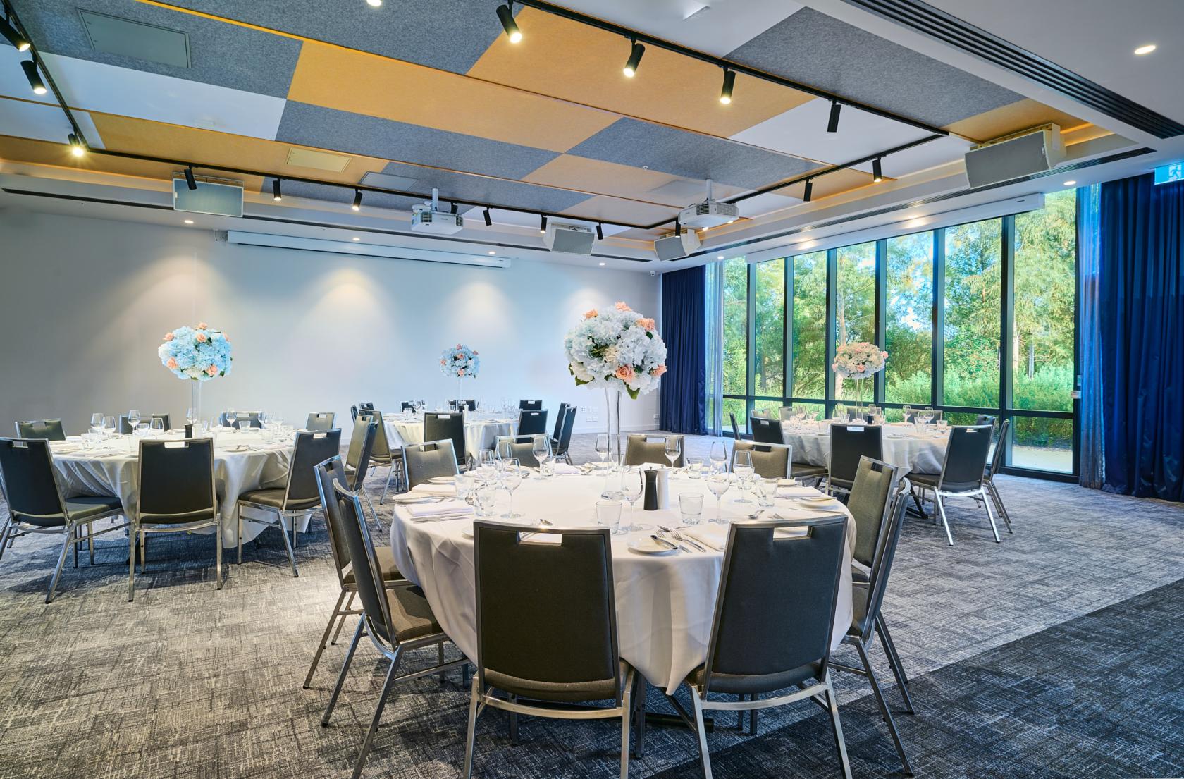 A hotel meeting room with round tables draped in white linens and floor-to-ceiling windows at Mantra Melbourne Epping.