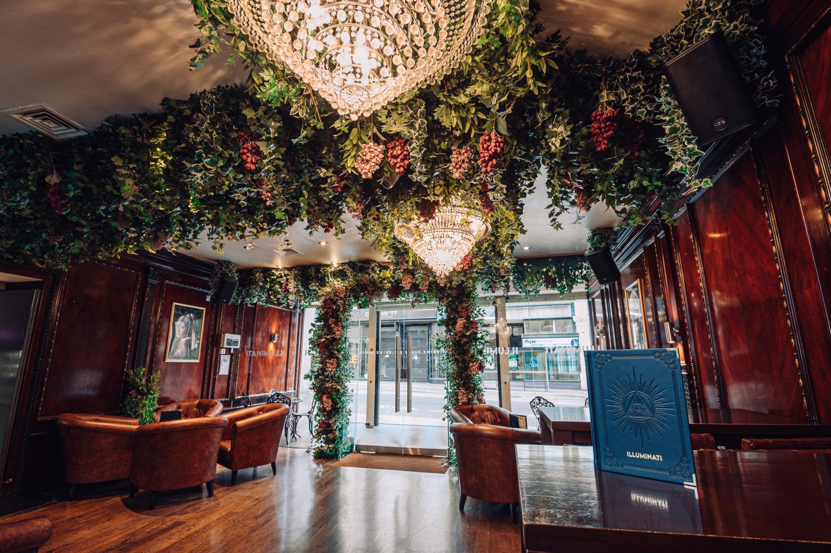 A cocktail bar in a nightclub with leather armchairs, wood paneling, and chandeliers under a leafy ceiling at Illuminati Bristol.
