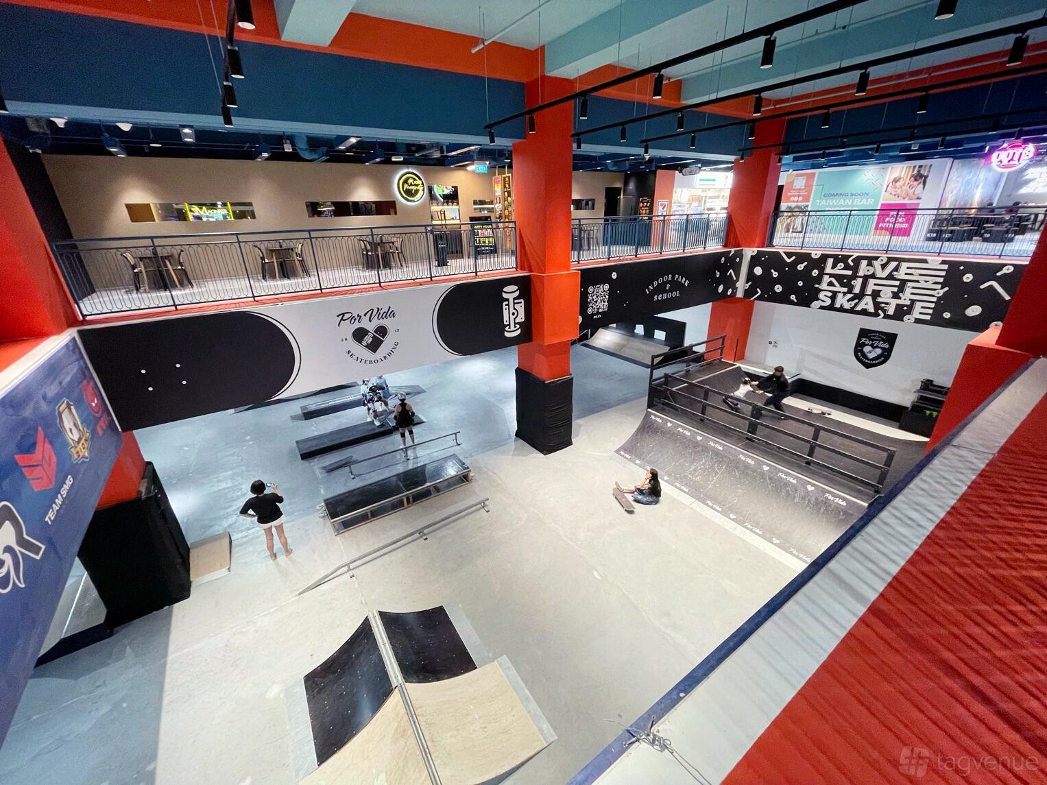 A sports hall with indoor skate ramps, graffiti-style wall art, and elevated lounge areas at Por Vida Skateboarding Indoor Park and School.