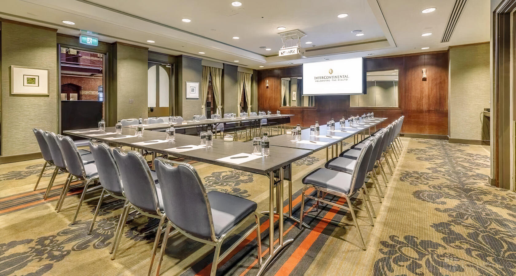 A hotel meeting room with U-shaped tables, cushioned chairs, patterned carpet, and a presentation screen at InterContinental Melbourne.