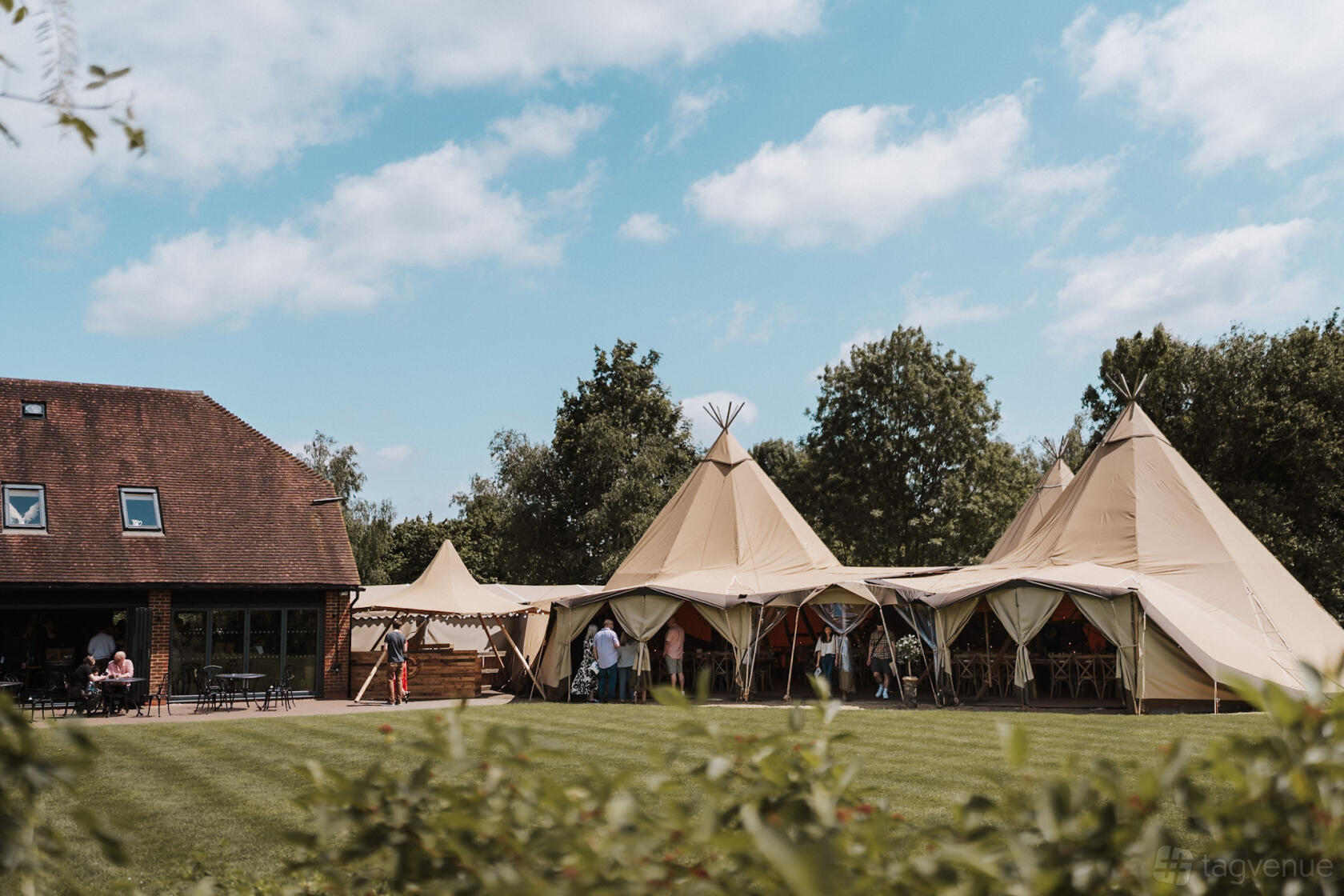 An outdoor event venue with tipi tents, lawn space, and a brick building surrounded by trees at The Gardens.