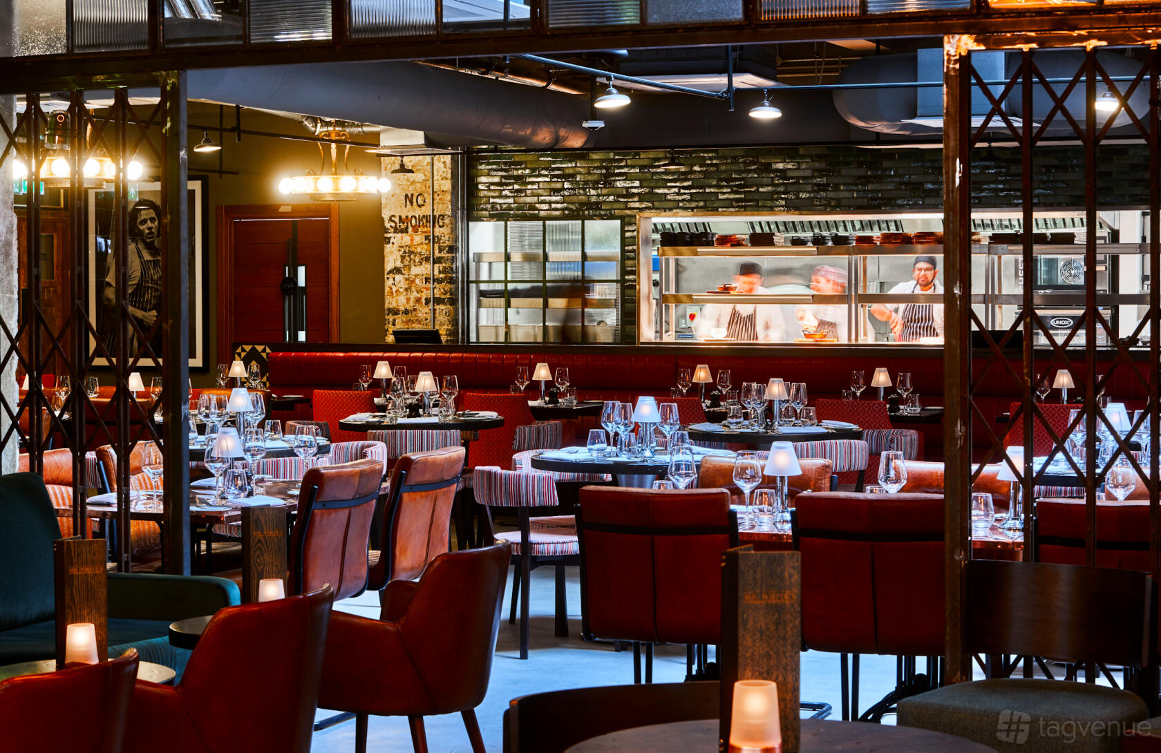 A restaurant with round tables set with glassware, red velvet chairs, and an open kitchen at New Road Hotel.
