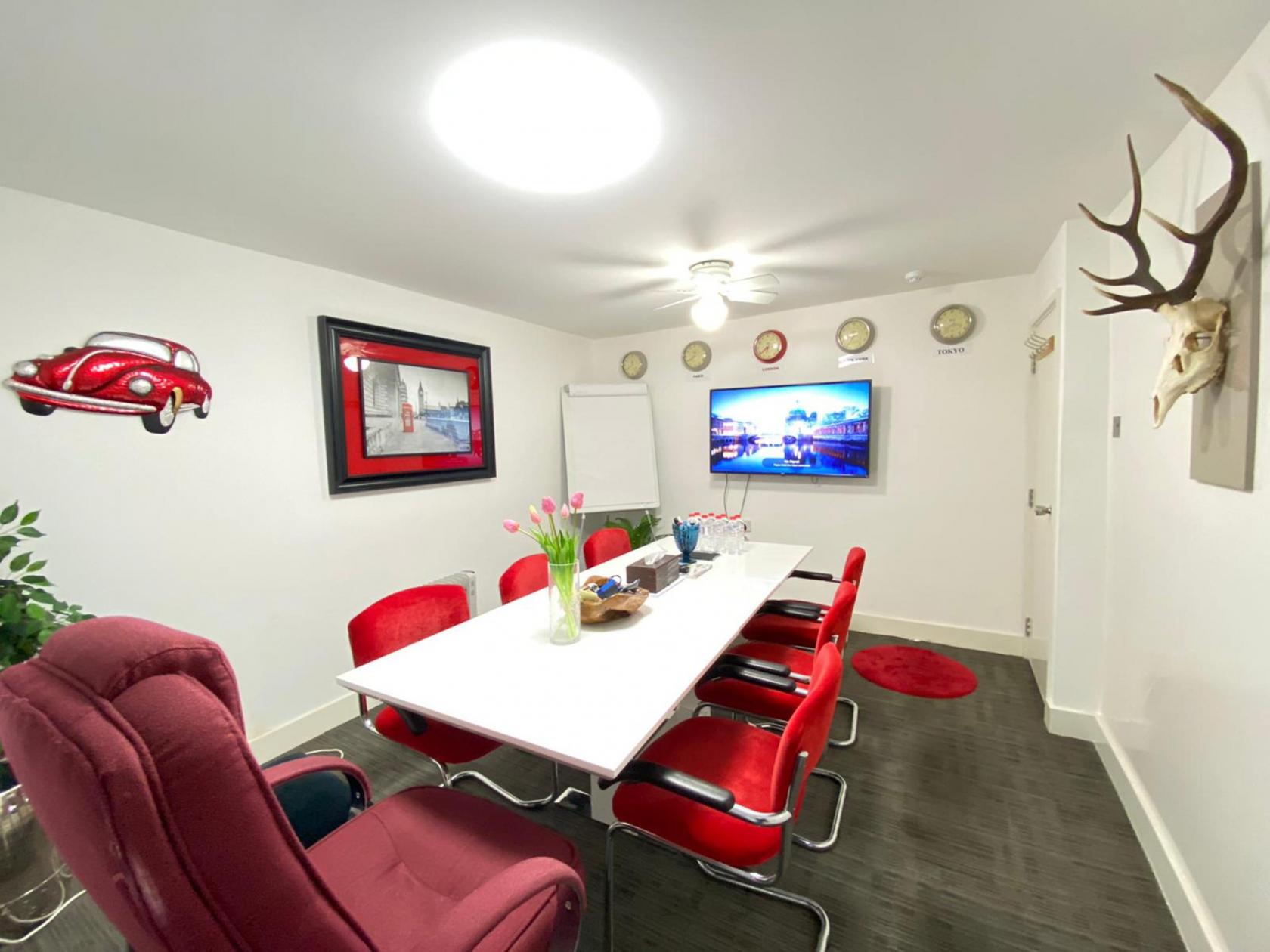 A meeting room with red chairs, a white table, wall clocks, and a mounted display screen at Meeting Room Brick Lane.