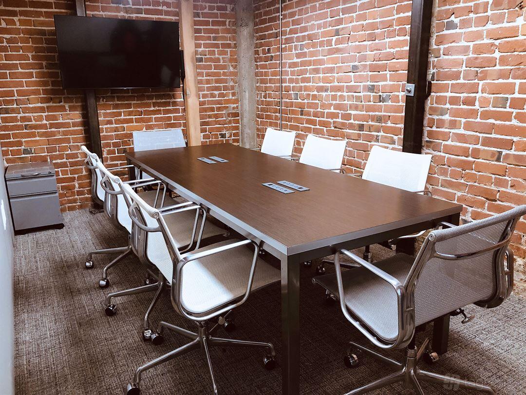 A meeting room with exposed brick walls, a dark rectangular table, and mesh office chairs at Temescal Works.