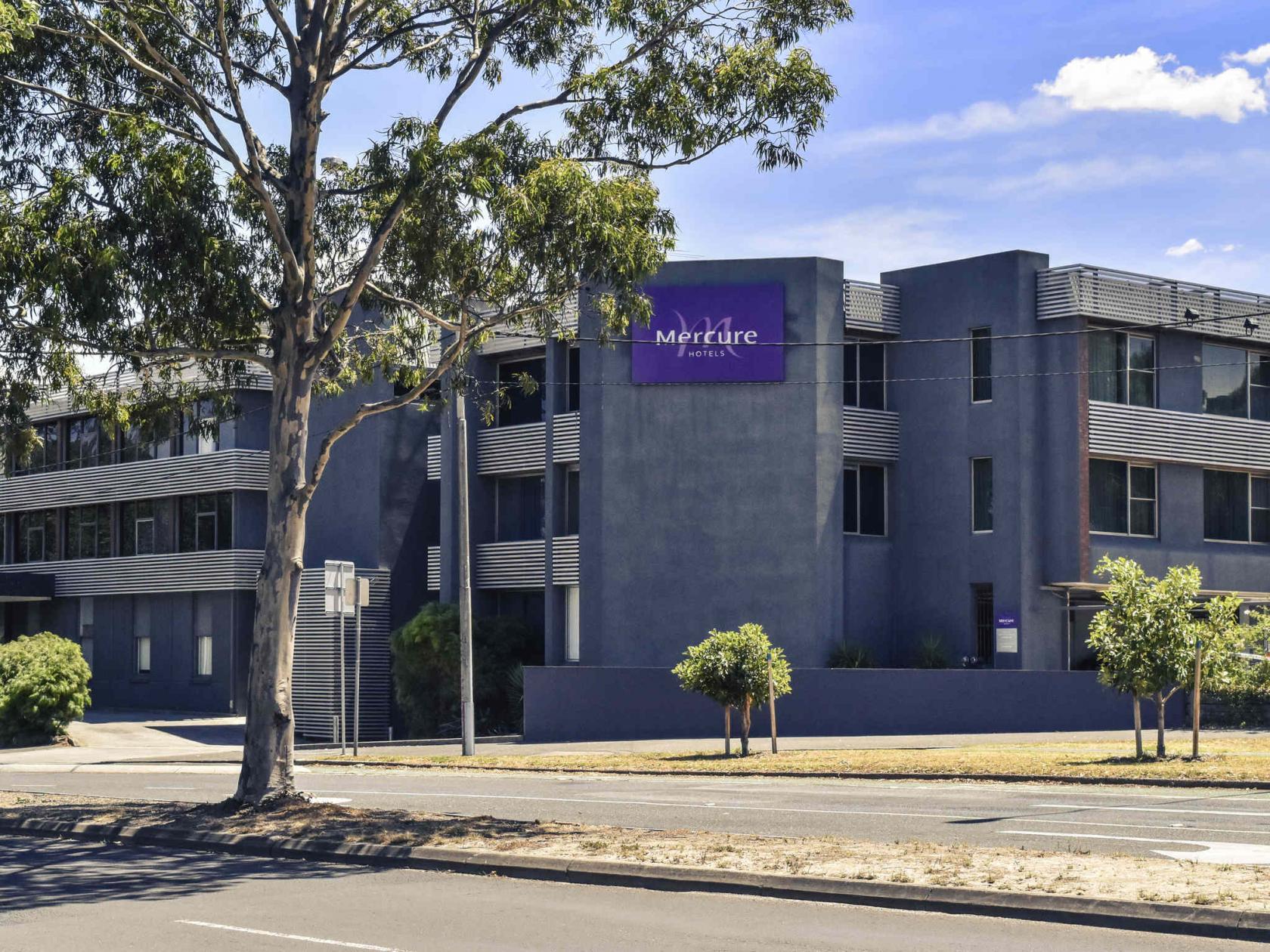 A hotel with a modern grey facade, large windows, and surrounding trees at Mercure North Melbourne.