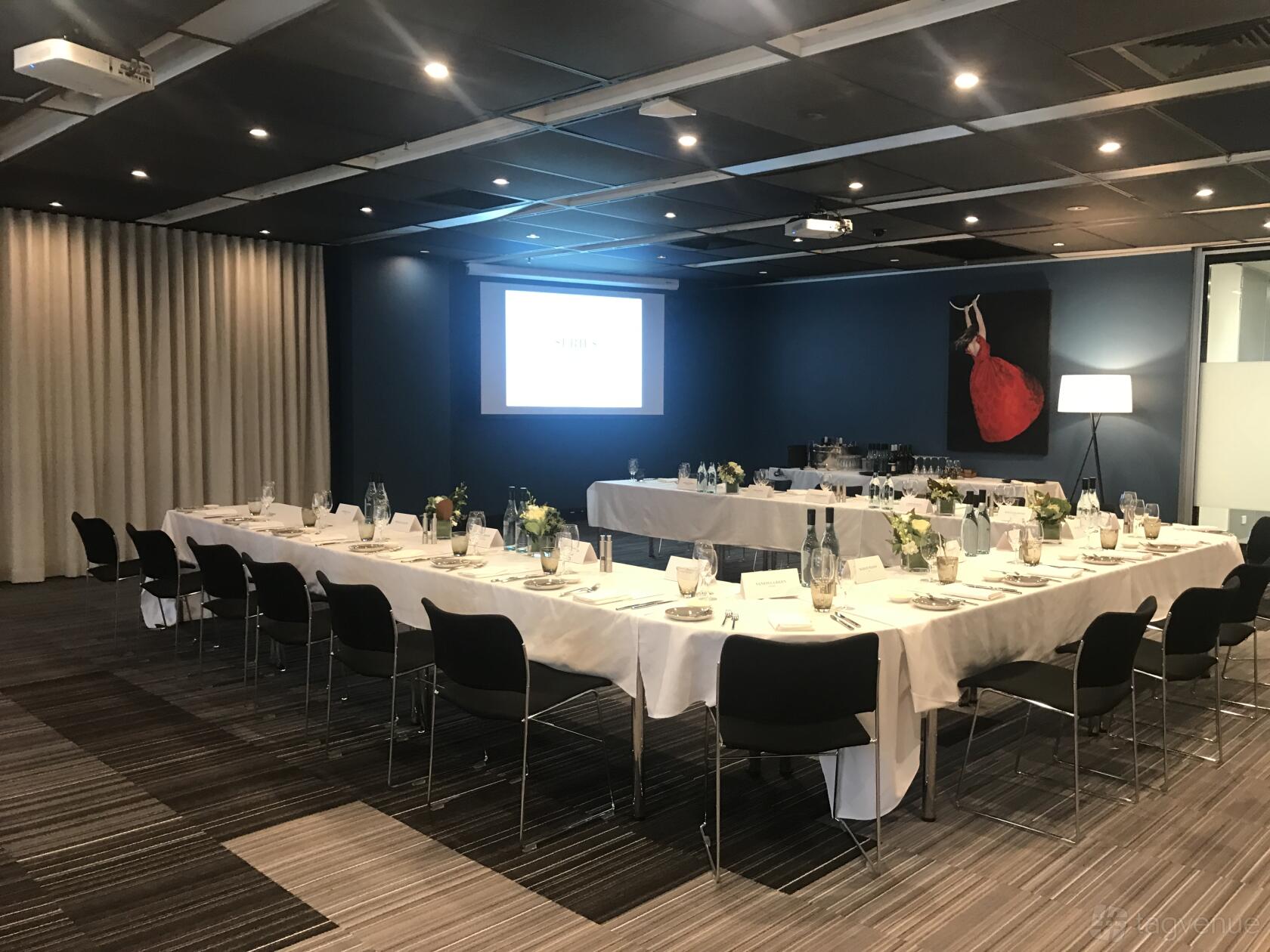 An event venue with U-shaped tables draped in white linens, a projector screen, and accent lighting at Cliftons Canberra – Moore St.