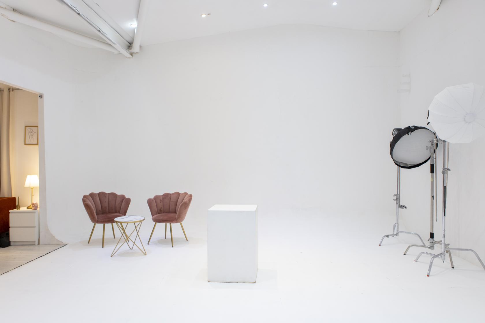 A photo studio with white walls, a pedestal, pink armchairs, and studio lighting equipment at Blankbox Studio.