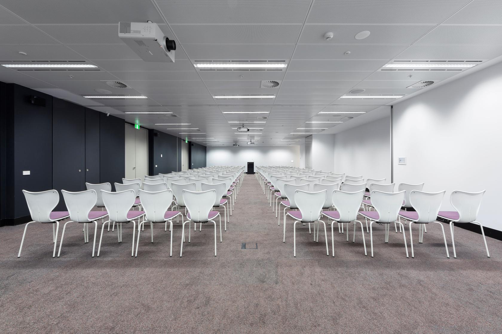 An event space with rows of white chairs facing a podium, carpeted floors, and ceiling-mounted projectors at Flex by ISPT at 4 National Circuit.
