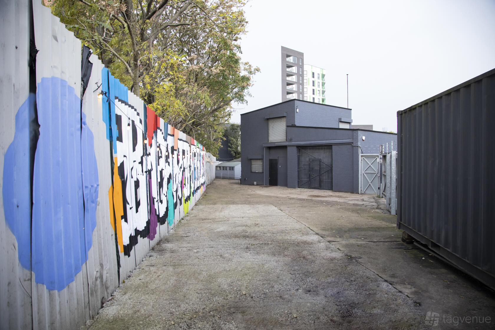 An outdoor courtyard with a graffiti-covered fence, concrete path, and industrial building at Bow Bunker.