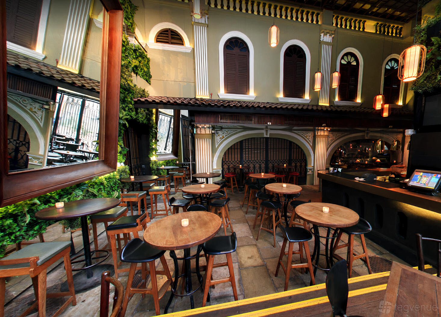 An outdoor bar area with high tables, wooden stools, hanging lanterns, and leafy greenery at Alley Bar.
