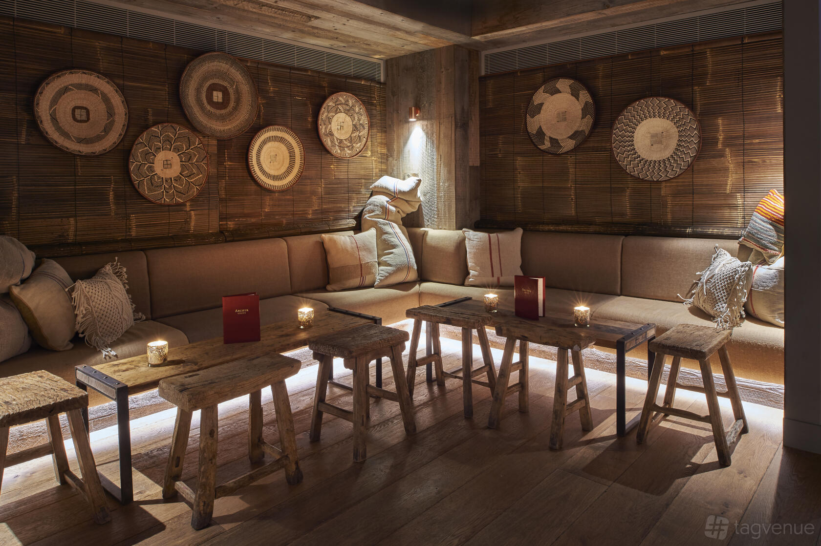 A cocktail bar lounge with wooden stools, built-in cushioned seating, soft candle lighting, and decorative wall baskets at Archer Street SW11.
