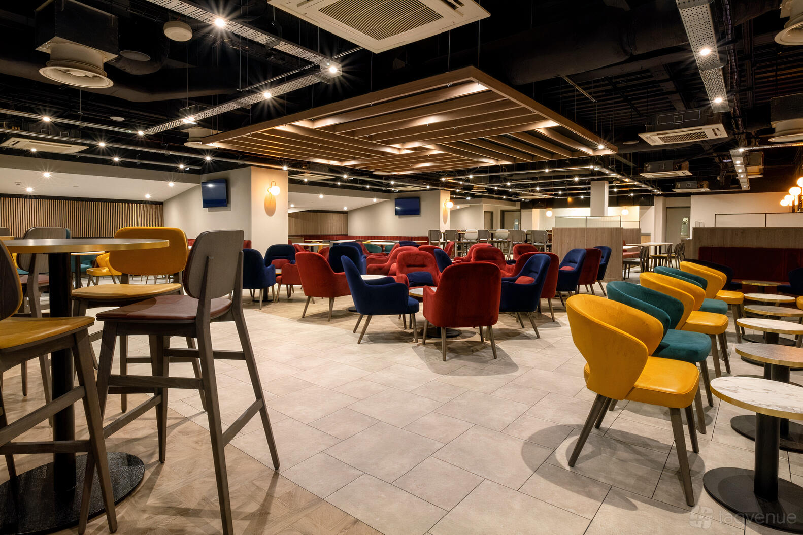 A lounge with colorful upholstered chairs and wooden tables under recessed lighting at Hampden Park Stadium.