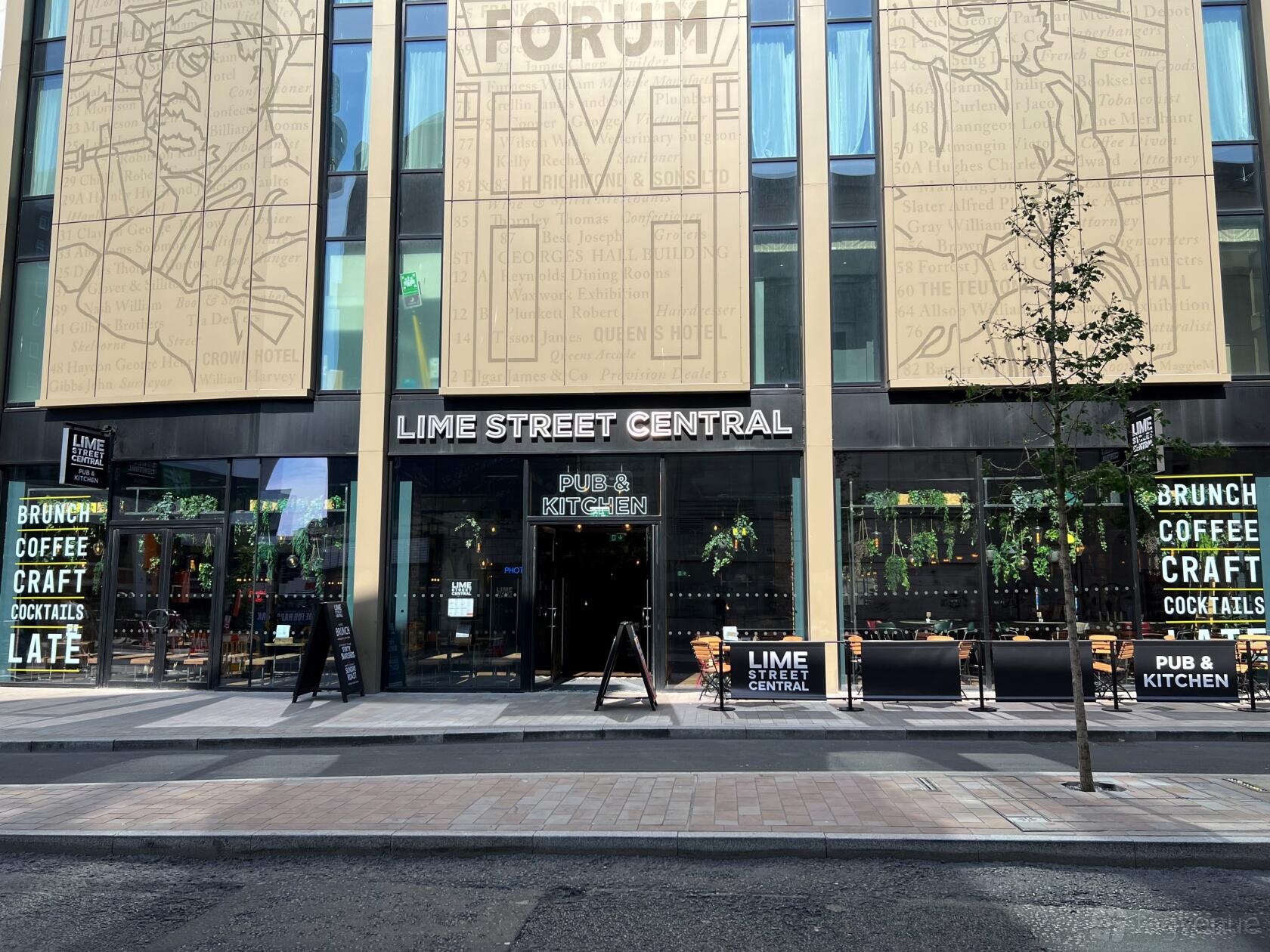 A bar with outdoor seating, large glass windows, and hanging signage at Lime Street Central.