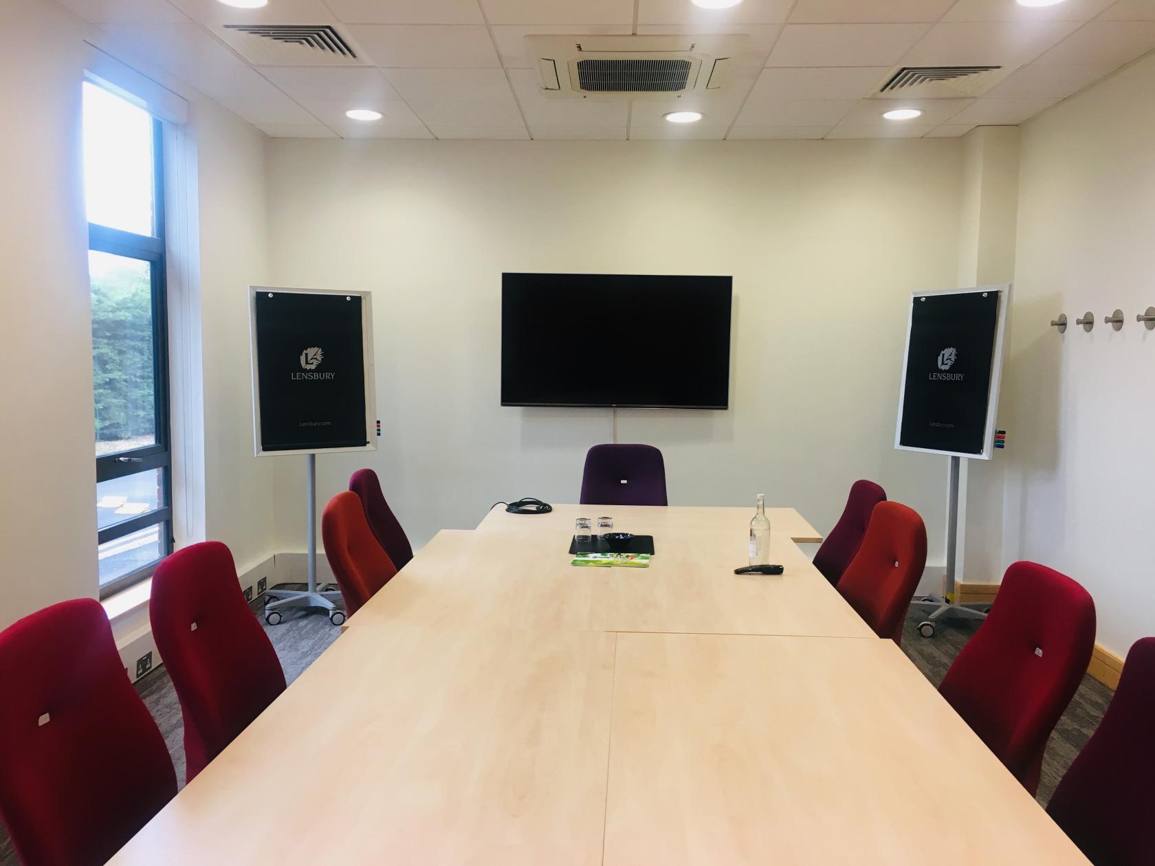 A hotel meeting room with a long rectangular table, red chairs, large wall-mounted screen, and natural lighting at The Lensbury.