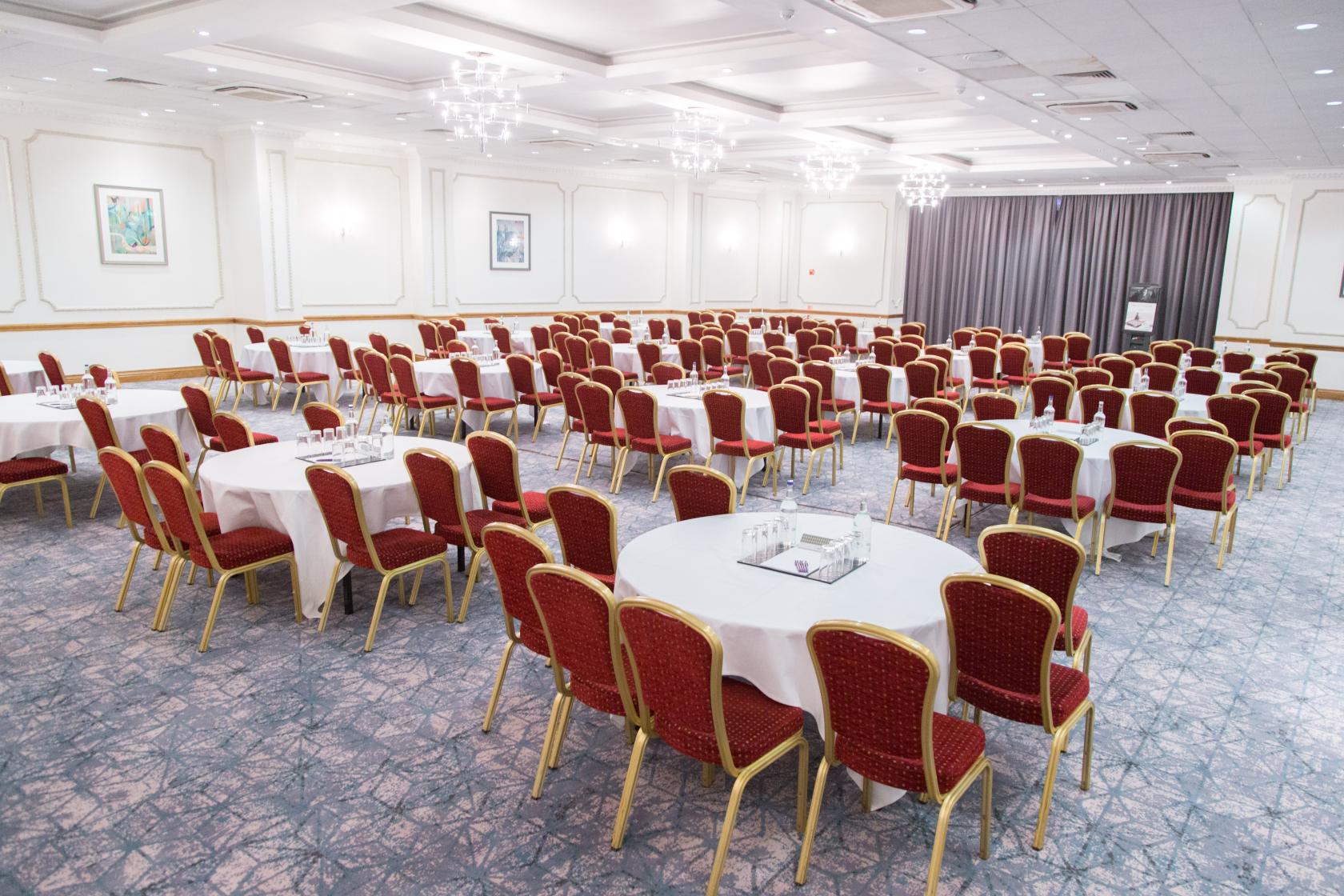 A banqueting hall with red cushioned chairs, round and rectangular tables, chandeliers, and framed artwork at Bristol Grand Hotel by Sunday.