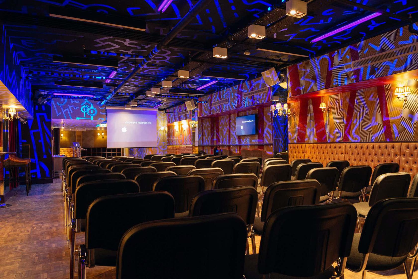 A function room with rows of black chairs, patterned blue lighting, and dual projector screens at Bounce Farringdon.