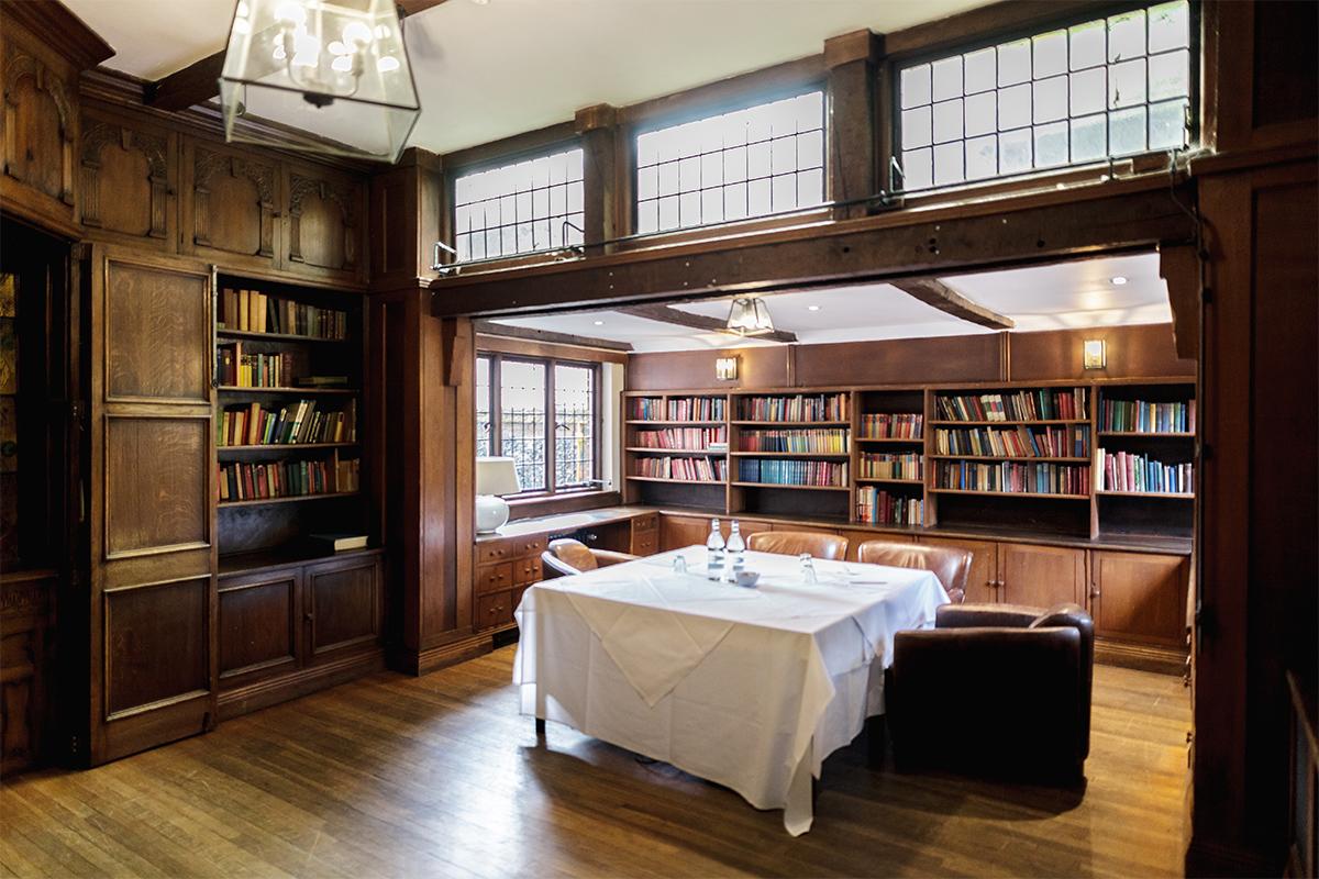 A hotel library with wood-paneled walls, floor-to-ceiling bookshelves, leather chairs, and a table with white linens at The Olde Bell.