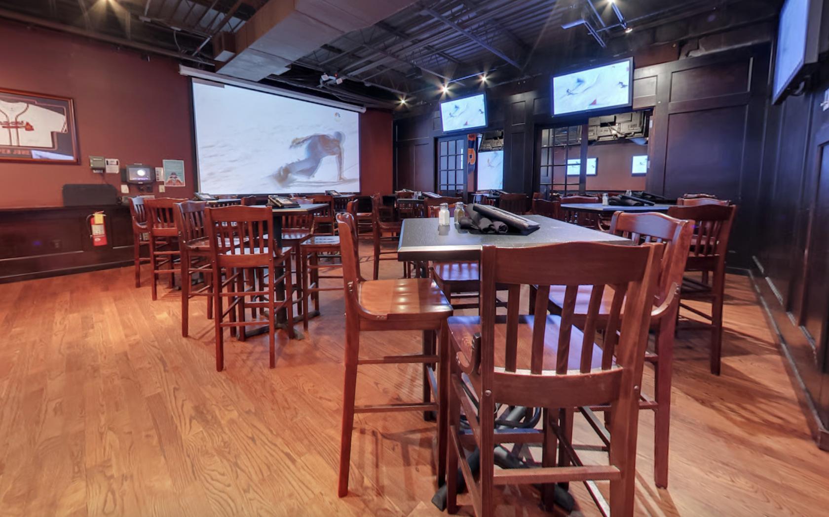 A dining room with wooden tables and chairs, multiple wall-mounted TVs, and sports decor at Hudson Grille - Sandy Springs.