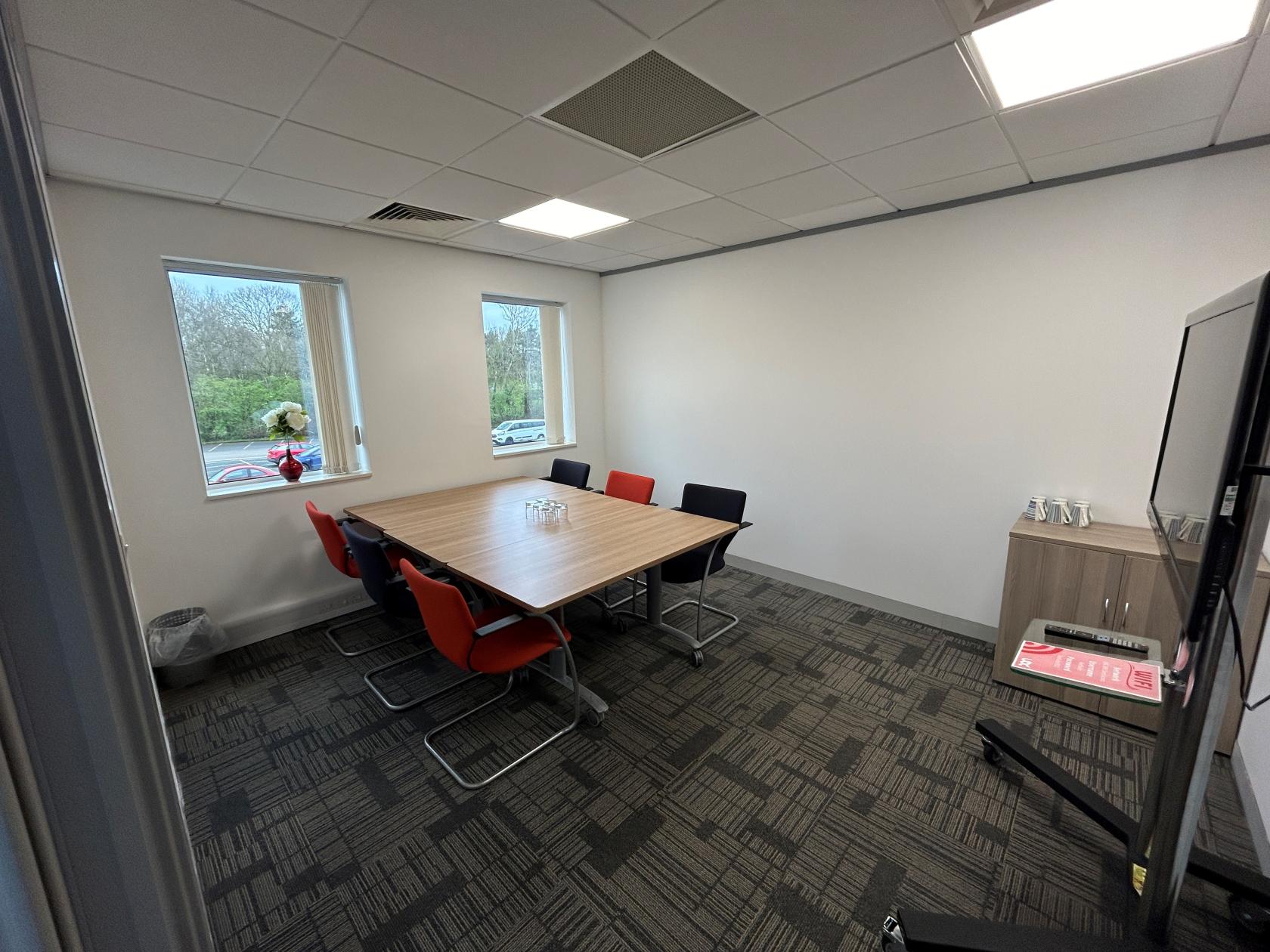 A meeting centre room with a rectangular table, mixed color chairs, large windows, and a wall-mounted screen at UBCUK Warrington at Rutherford House.