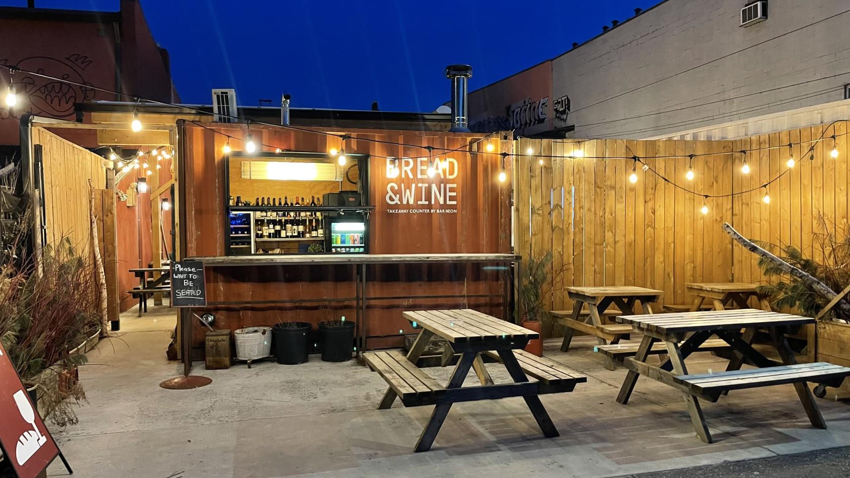 An outdoor terrace with wooden picnic tables, string lights, and a walk-up bar at Bar Neon.