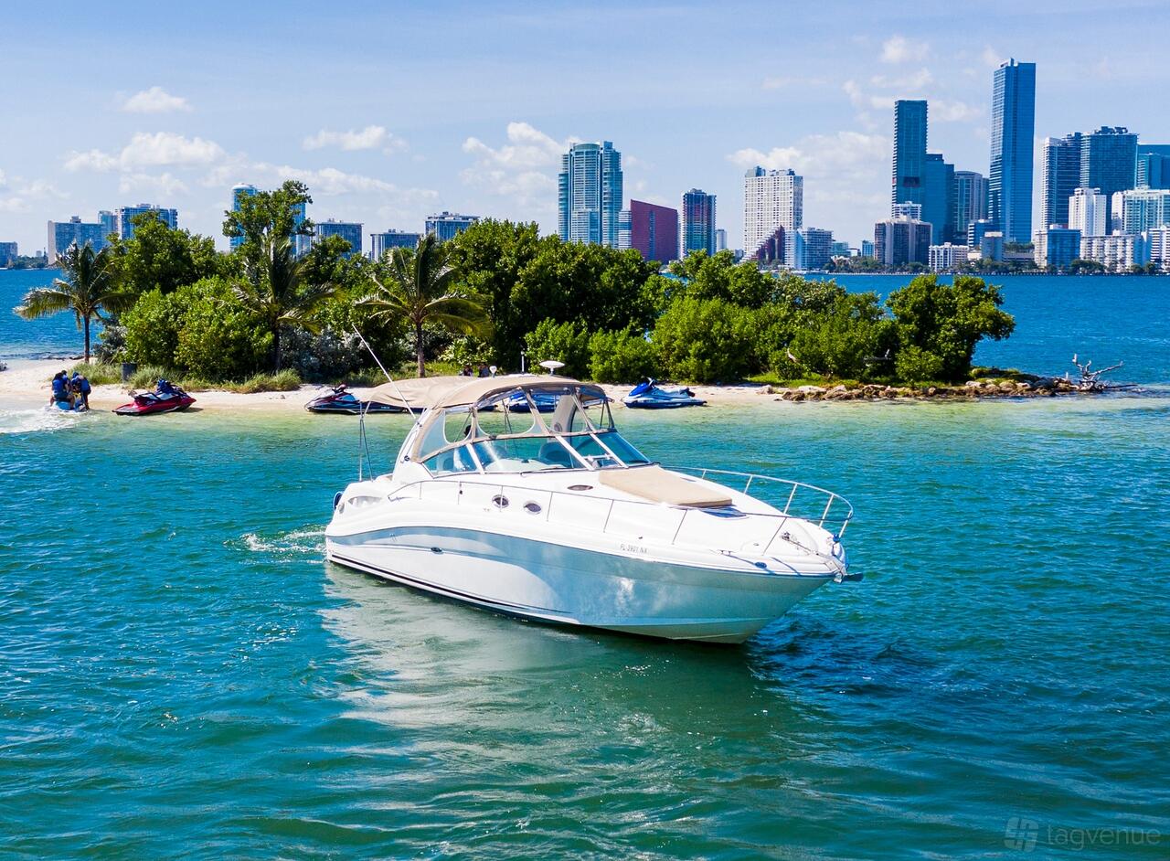 A white motor yacht with a tan canopy cruising near a small island and city skyline at Miami Like a Boss @ Alton Rd.