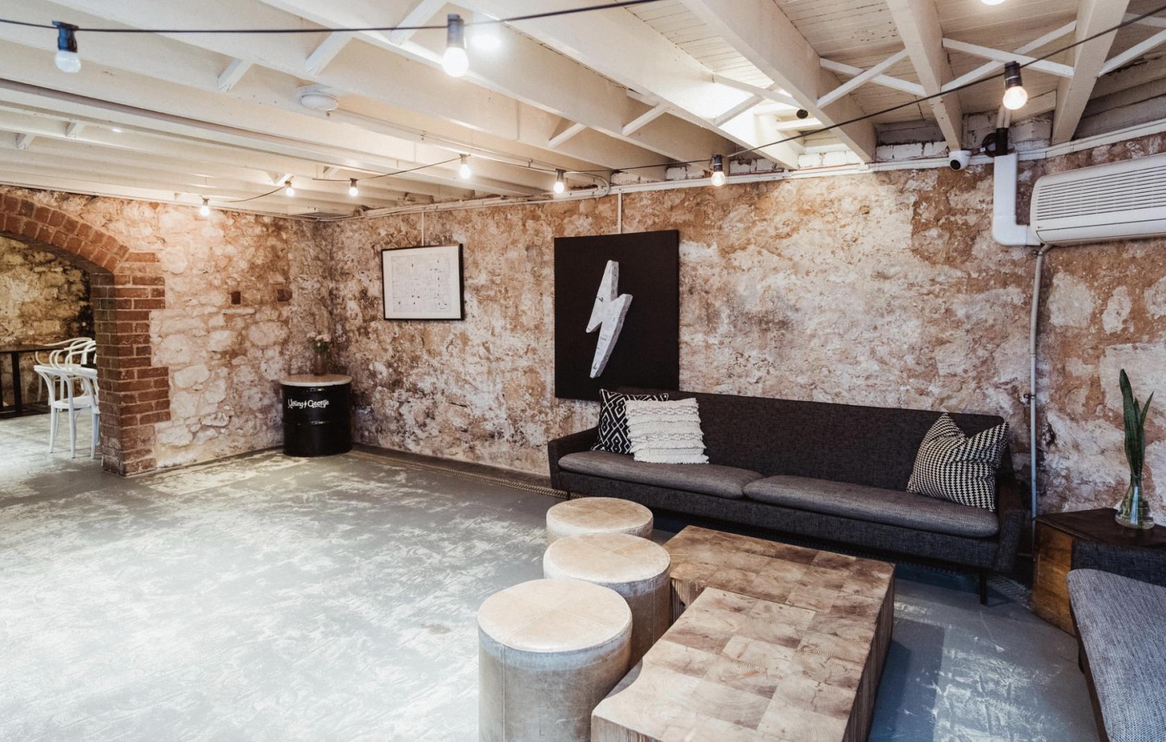 A bar with exposed stone walls, string lights, and a lounge area with a dark sofa in the Cellar at Young George.