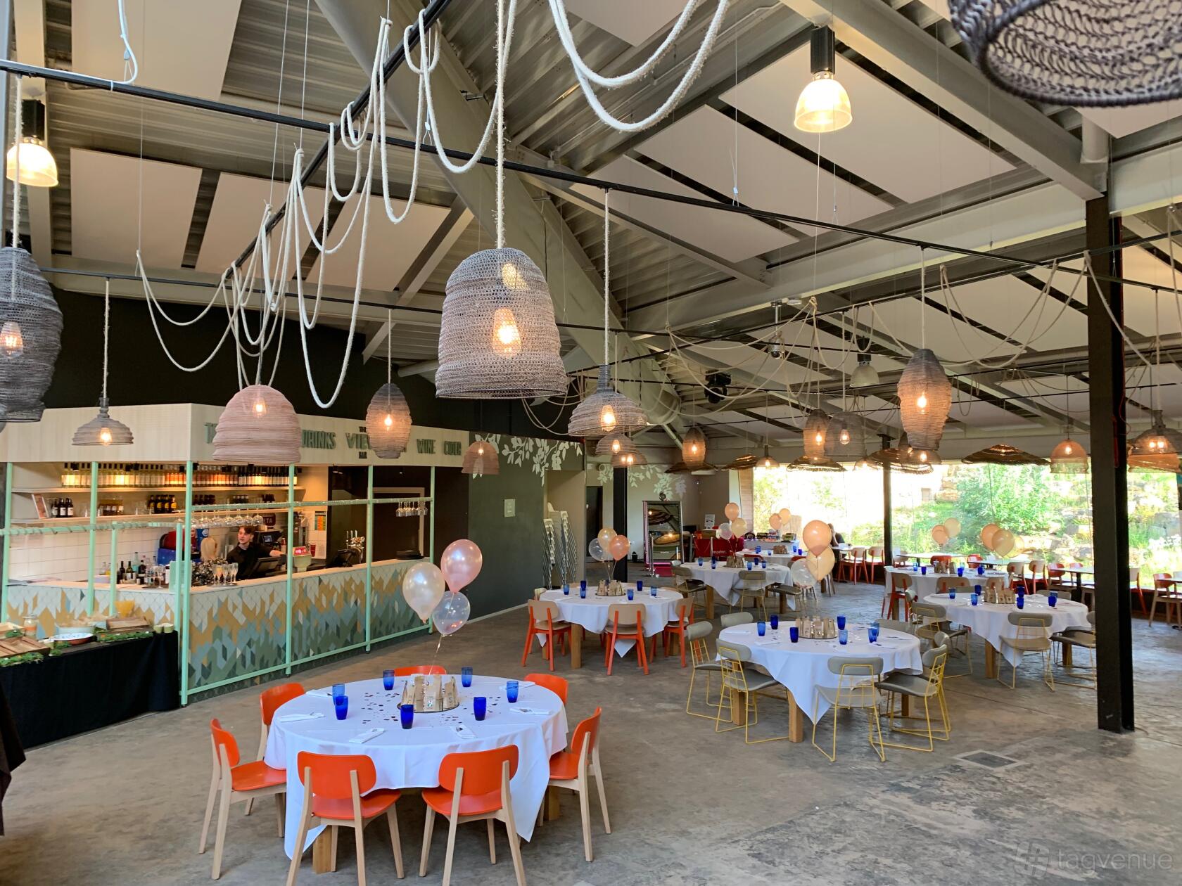 A spacious hall with round tables, orange chairs, hanging woven pendant lights, and a bar area at Twycross Zoo Himalaya Room.