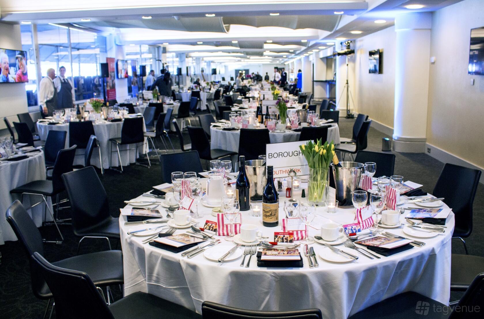 A stadium suite with round tables set with glassware, wine bottles, and fresh flowers at Chelsea Football Club.