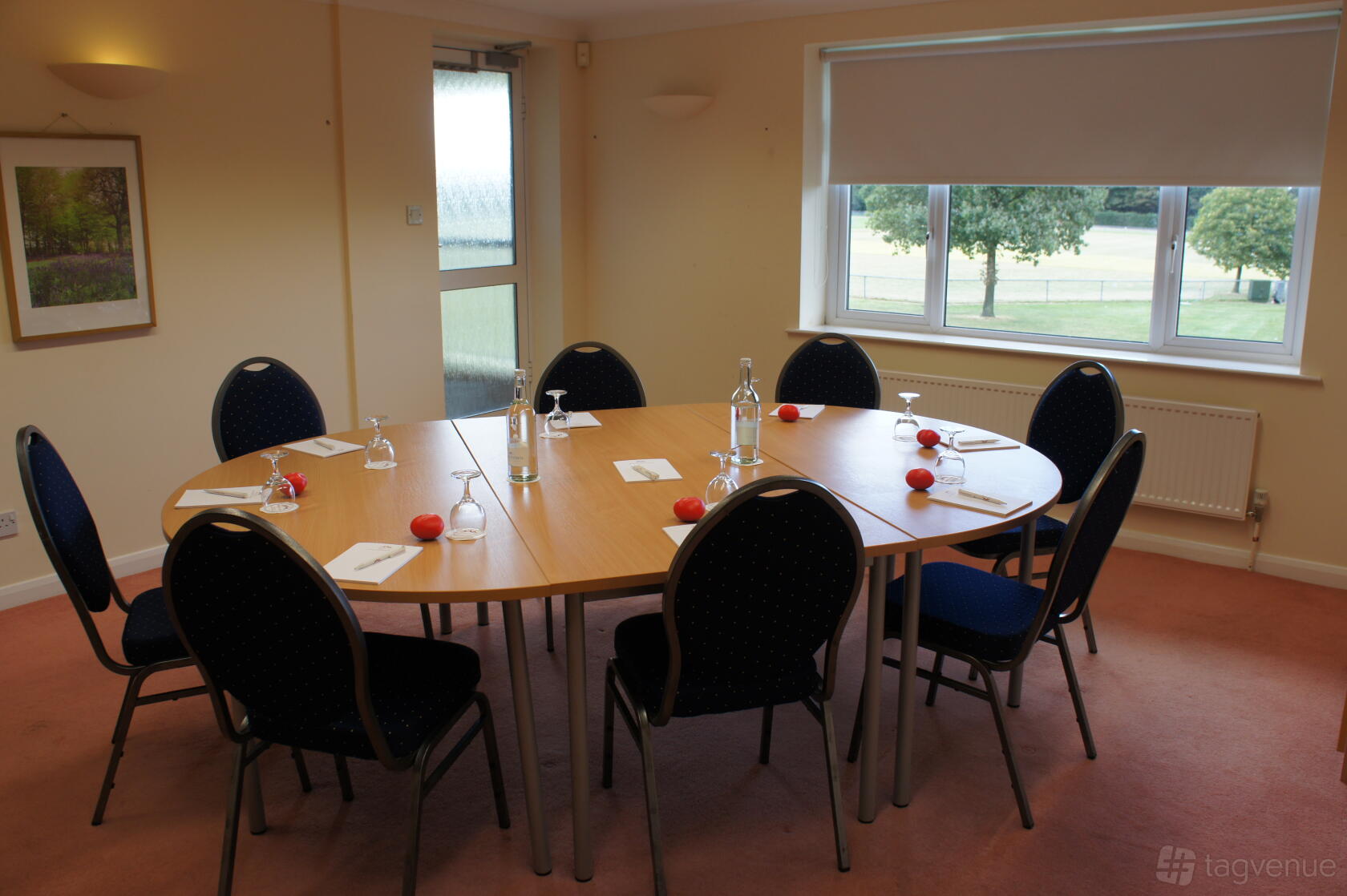 A meeting room with a round table, padded chairs, bottled water, and large window overlooking trees at Kent Event Centre.