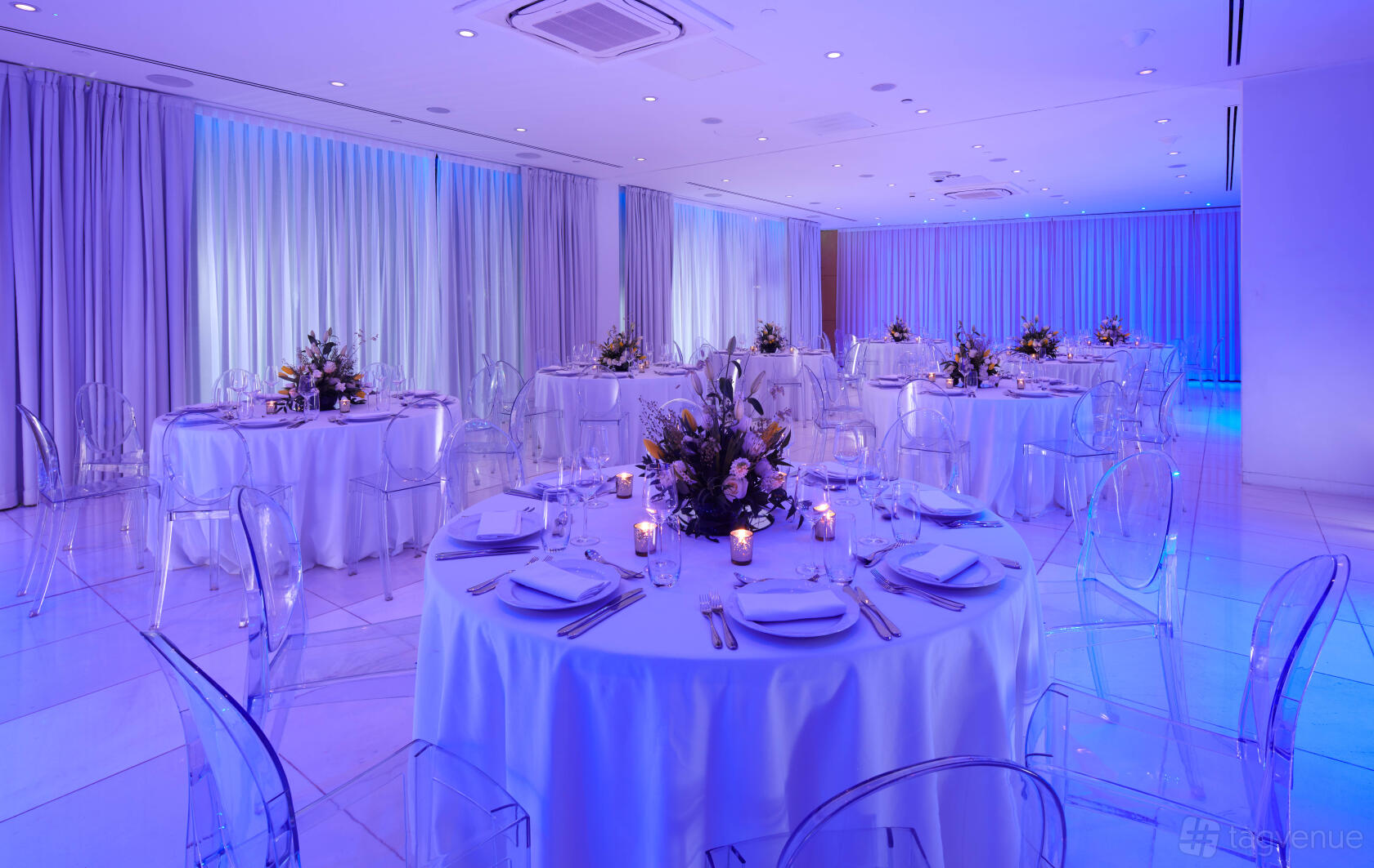 An event space with round tables draped in white linens, elegant place settings, and purple ambient lighting at St Martins Lane London.