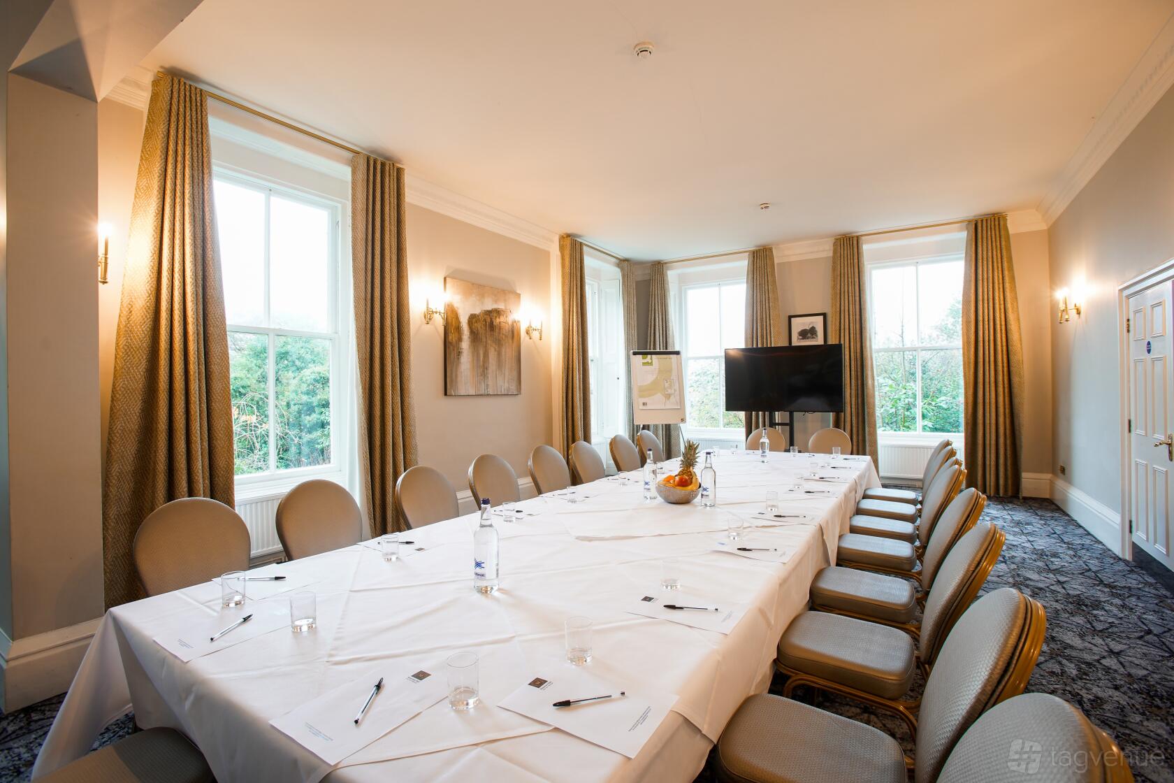 A meeting room with large windows, neutral walls, and a long U-shaped table with chairs at Makeney Hall Hotel.