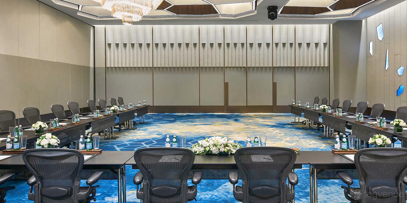 An event space in a hotel with a geometric patterned blue carpet, chandeliers, and U-shaped meeting tables at Pan Pacific Singapore.