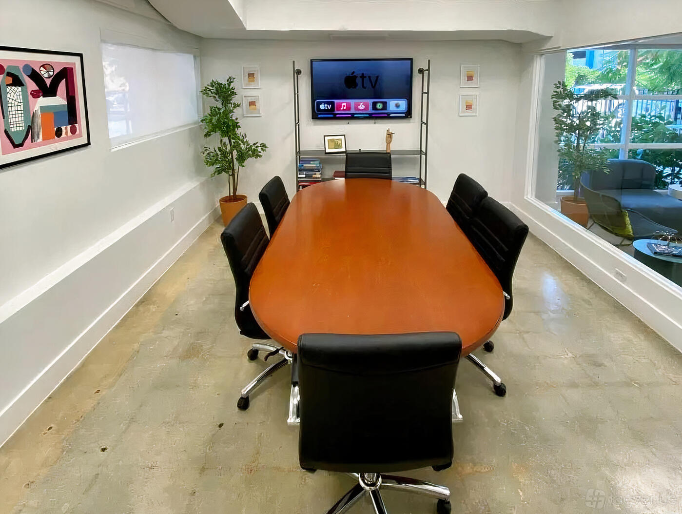 A coworking meeting room with a long oval table, black chairs, wall-mounted TV, and large windows at The Div Project.