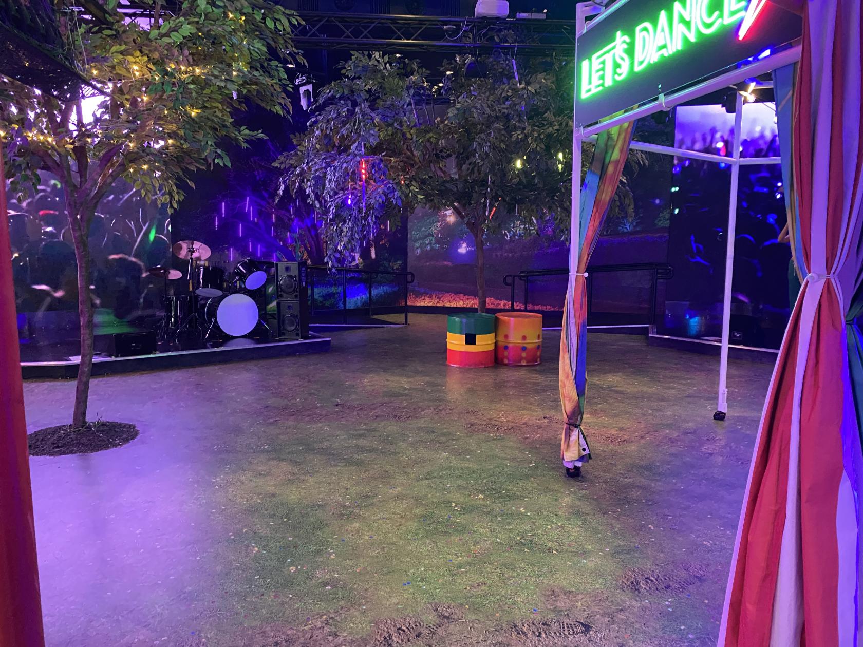 An event space with artificial trees, colorful barrel seating, stage lighting, and a neon 'Let’s Dance' sign at Madame Tussauds.