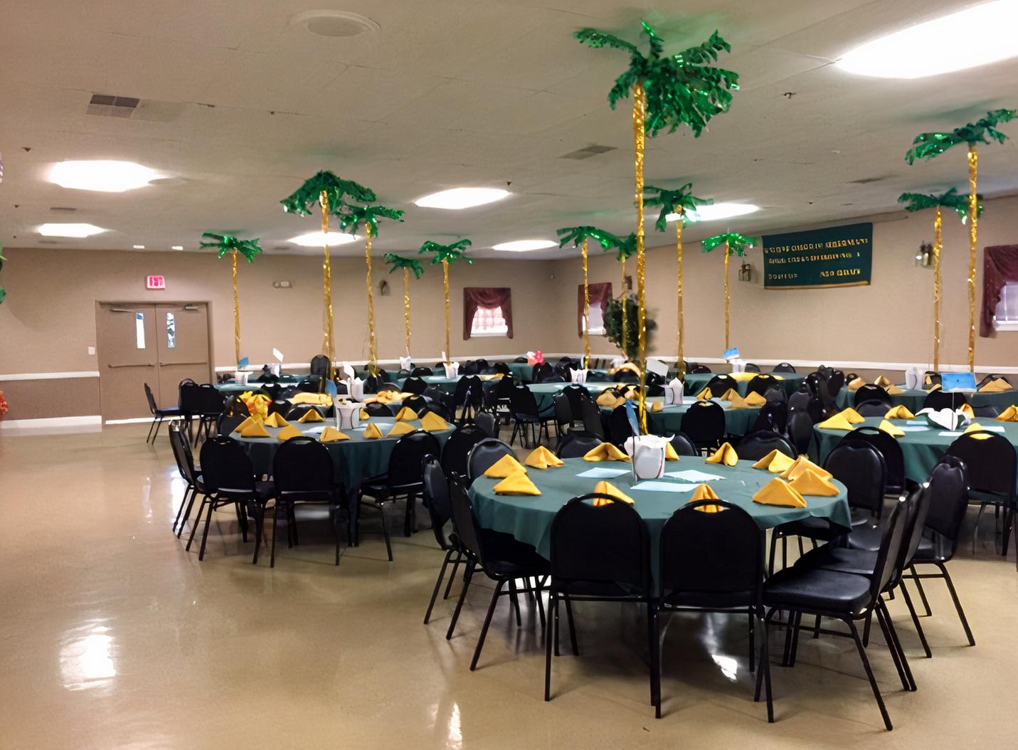 An event venue with round tables dressed in green tablecloths and gold napkins, featuring artificial palm tree centerpieces at Hibernian Club.