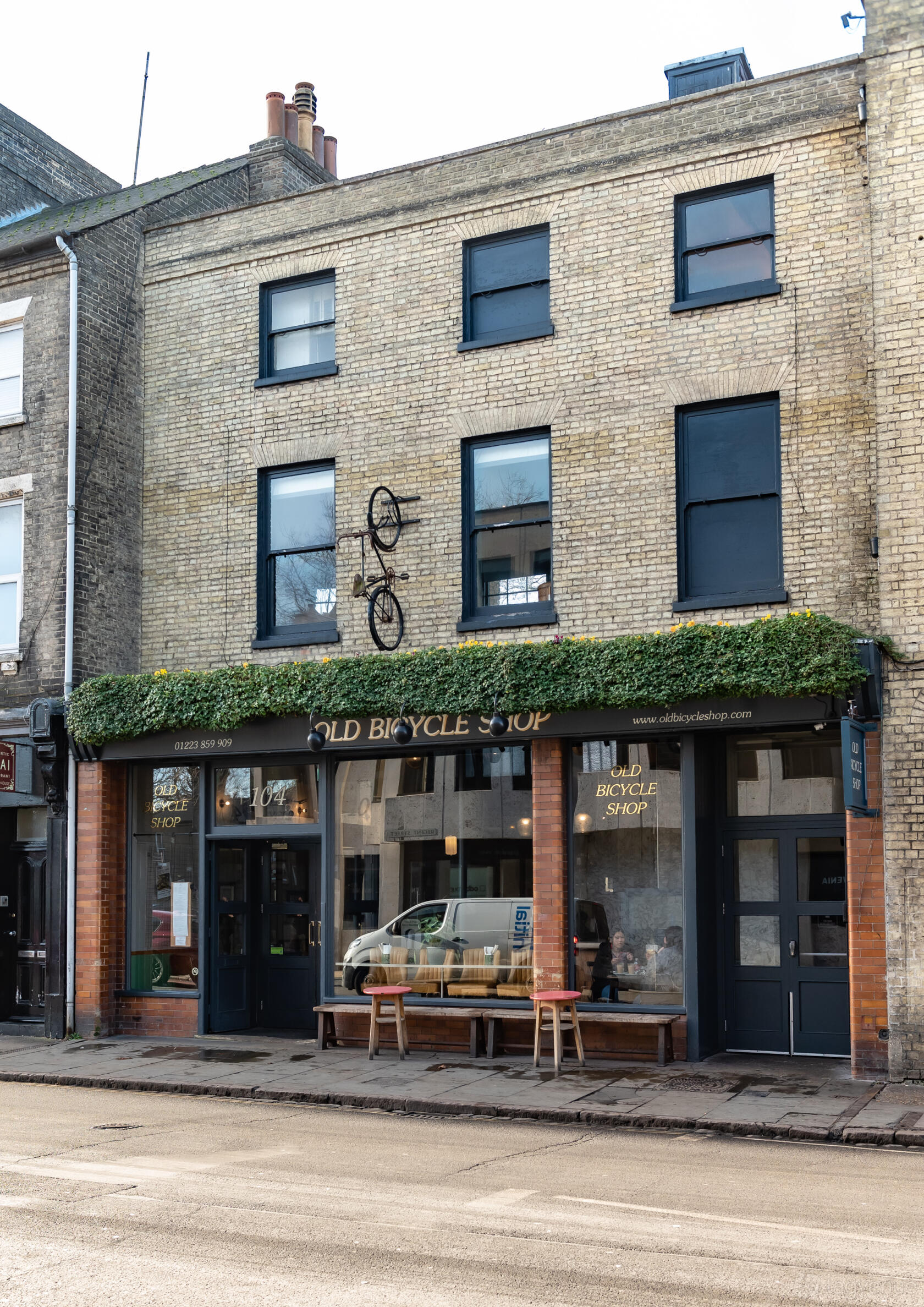 A pub with ivy-covered brick exterior, large windows, and a bicycle mounted above the entrance at Old Bicycle Shop.