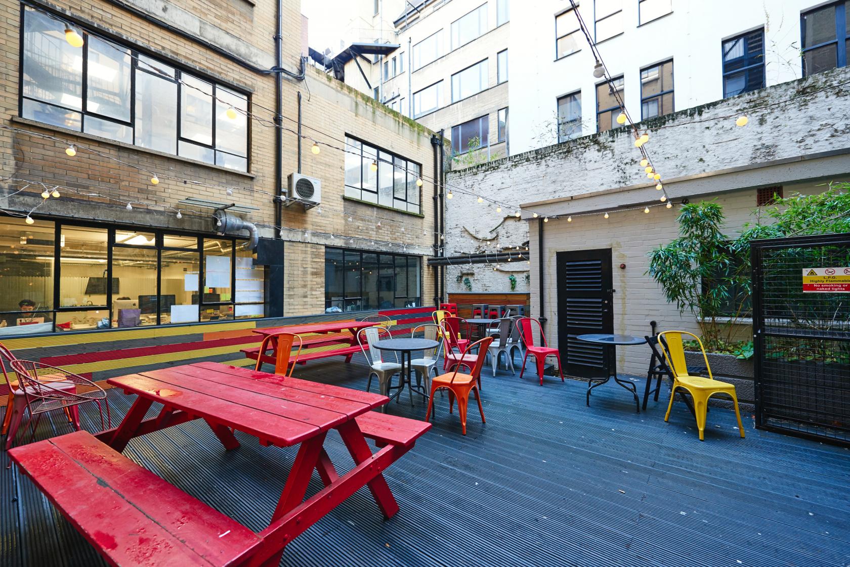 An outdoor coworking space with red picnic tables, yellow chairs, string lights, and urban building views at The Worker's League.