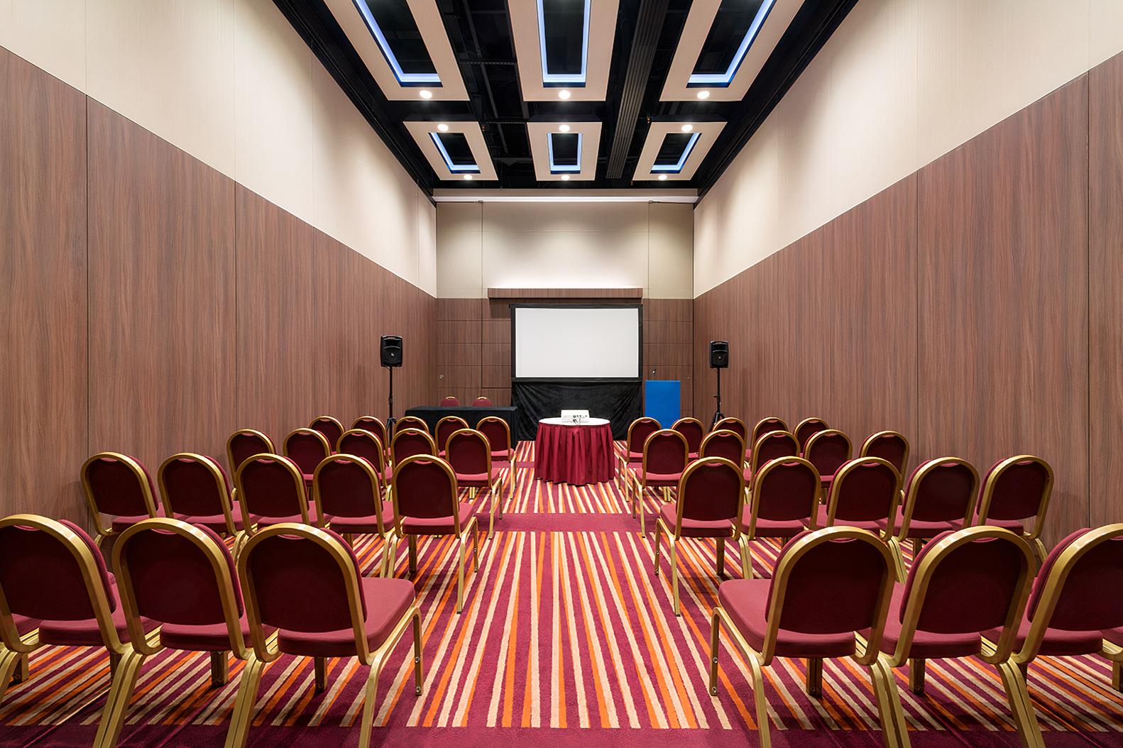 A meeting room with rows of red chairs, striped carpet, and a projector screen at Mercure London Earl's Court.