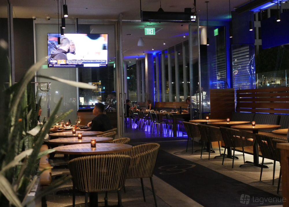 A restaurant with round wooden tables, wicker chairs, candlelit centerpieces, and blue accent lighting at Pita Bar + Grill.