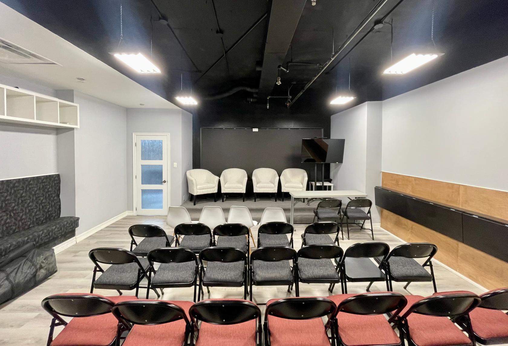 An event venue with a small stage, white armchairs, black folding chairs, and overhead lighting at Entrepreneurs Point.