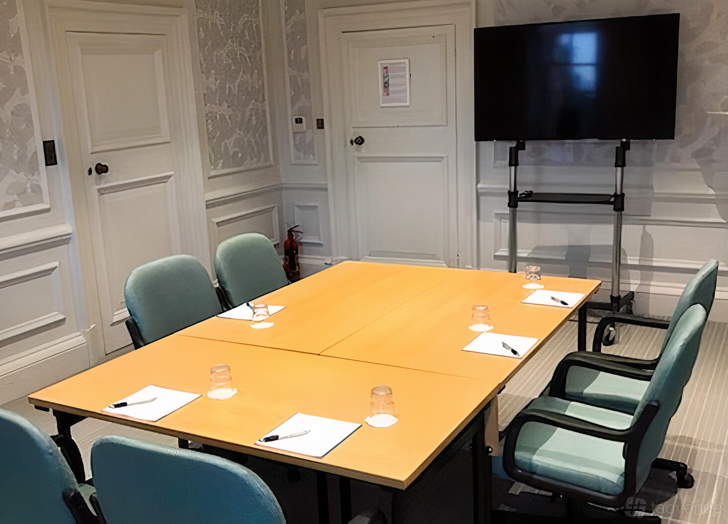 A hotel meeting room with a square conference table, blue chairs, stationery, and a large TV screen at Barnett Hill Hotel.