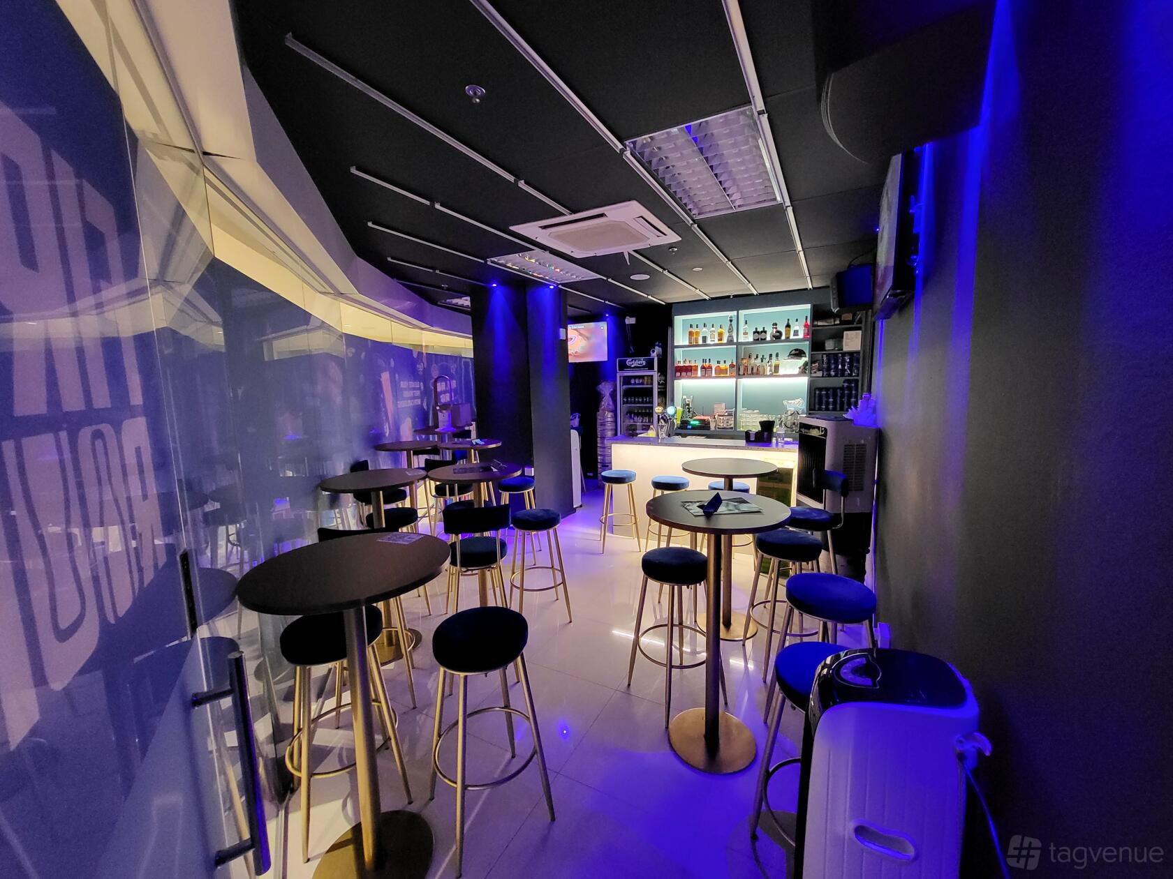 A karaoke bar with a lit-up bar counter, high round tables, and black bar stools at First Round.
