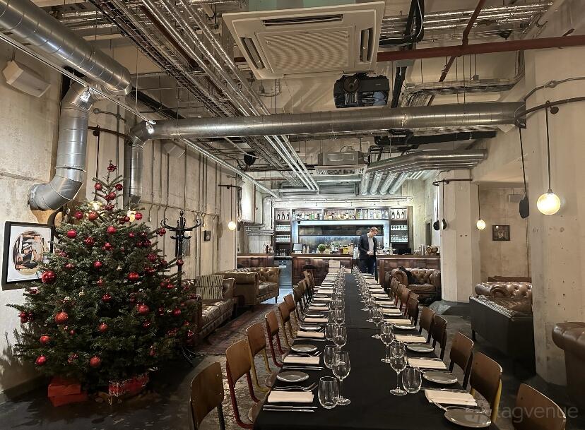 A lounge with a long banquet table set for dining, exposed pipes, and a decorated Christmas tree at Bread Street Kitchen, Southwark.
