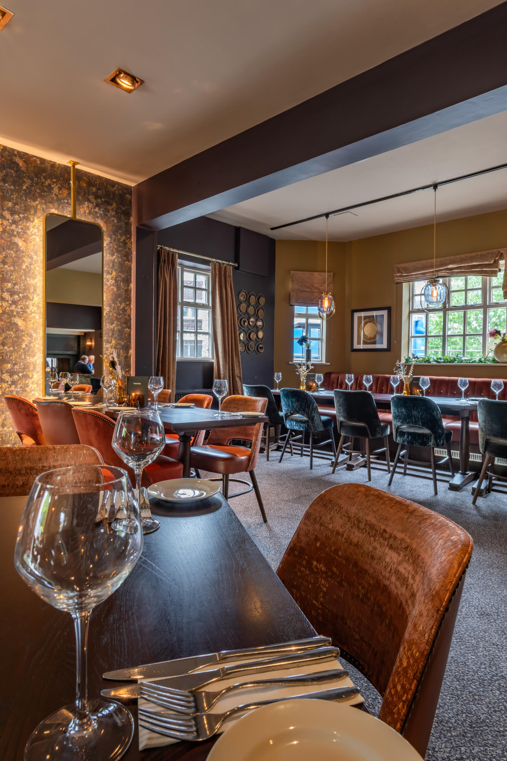 A dining room in a pub with velvet chairs, set tables, pendant lights, and large windows at The Oat Sheaf.