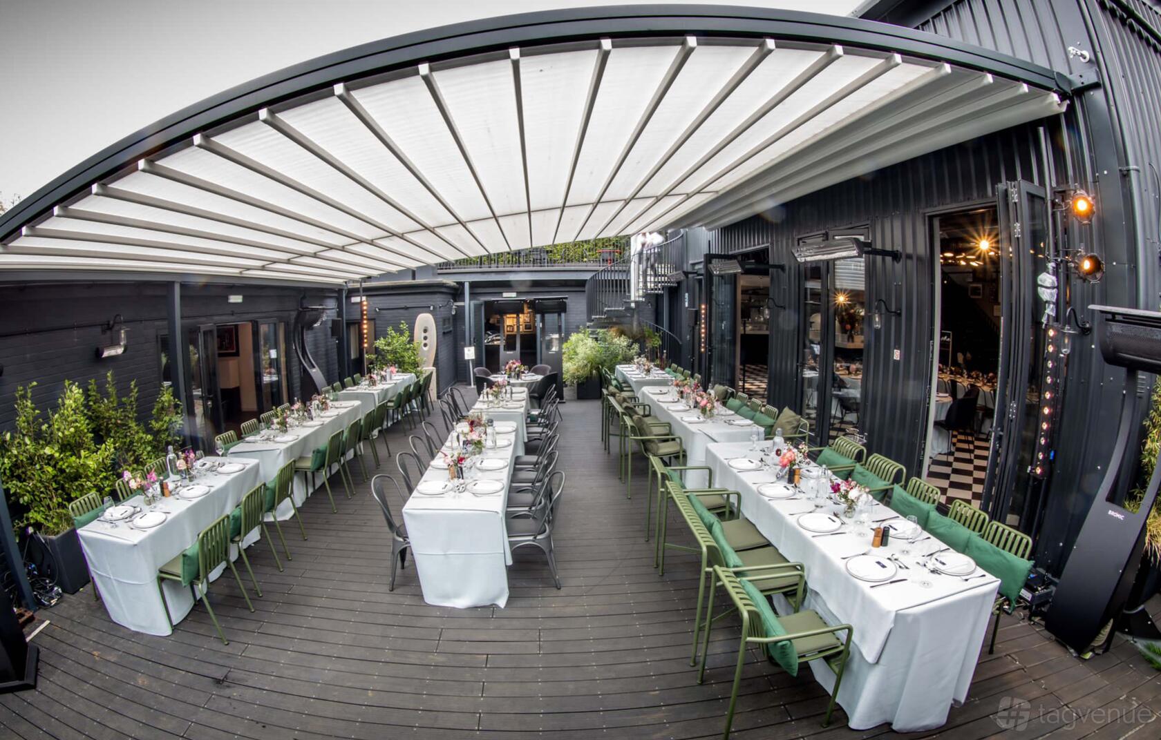 An outdoor dining area with long tables covered in white linens and green chairs at The Engine Rooms Events Space.