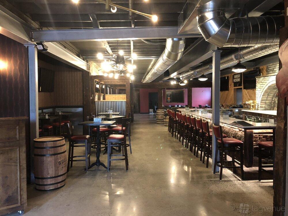A restaurant with polished concrete floors, exposed ductwork, and a long bar lined with high chairs at The Oakley - Kitchen & Cocktails.
