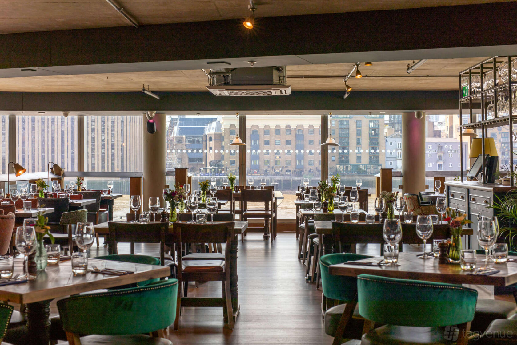 A lounge in a pub with large windows, wooden tables set with glassware, and views of the city at The Oyster Shed.