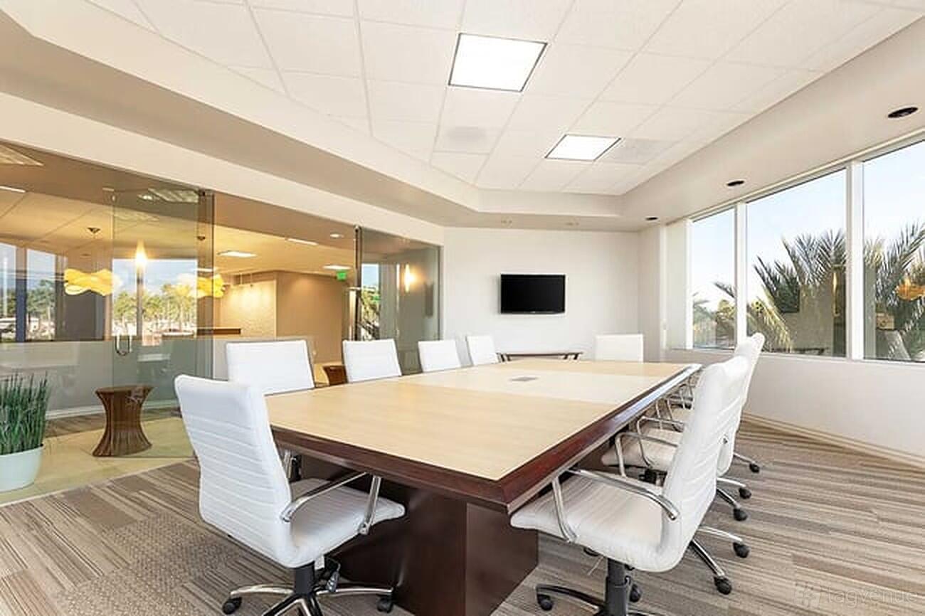 A meeting room with a large rectangular table, white chairs, floor-to-ceiling windows, and a wall-mounted screen at Premier Workspaces - Foothill Ranch.