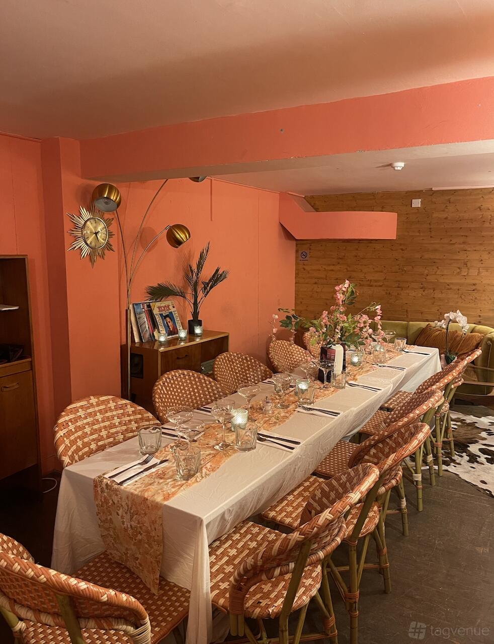 A lounge in a cocktail bar with coral walls, a long table with floral arrangements, and woven chairs at Studio48 Bar & Kitchen.