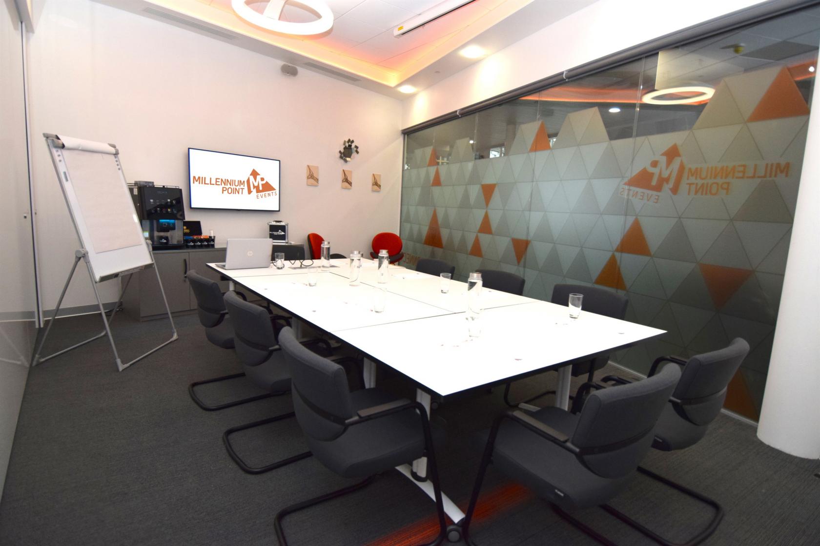 A meeting room with a long white conference table, black chairs, glass partition, and presentation screen at Millennium Point.